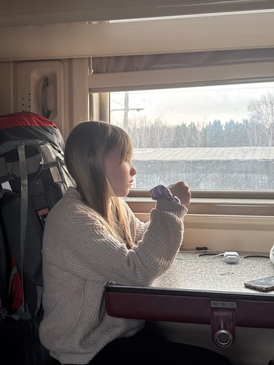 A young female is depicted seated inside a train compartment, gazing out a window at a landscape consistent with travel through the vicinity of Usolye-Sibirskoye, Russia. The subject, appearing to be a young adult, has long, straight, light brown hair and is wearing a light beige, chunky knit sweater over dark trousers. A light purple scrunchie is visible on her left wrist. Her left arm rests on a light grey, speckled table with a dark red upholstered edge, where a white Apple AirPods charging case, a white charging cable, a dark-colored smartphone with a transparent or glitter-flecked case, and a small metallic ring are present. To the left of the female, a large dark grey and red hiking backpack, bearing a "SNOVA TOUR" label, occupies a seat or storage space. The train's interior features light-colored paneling and a cream-colored fixture with a receiver-like handle and perforations. The exterior view through the window shows a pale, overcast sky above a wintery or early spring landscape. This landscape includes bare deciduous trees in the foreground, darker evergreen trees behind them, and a low-lying building with a corrugated roof in the mid-distance. A utility pole with crossbars is partially visible on the left side of the window frame. Natural light from the window illuminates the scene.