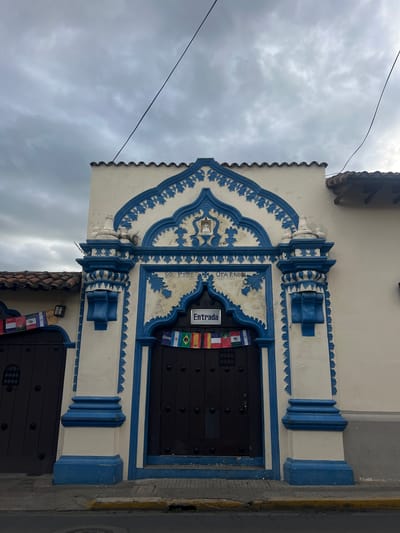 The image shows the facade of a building in León, Nicaragua, with ornate blue and white architectural details. The building has a large, dark wooden door that serves as the entrance. Above the door, a sign reads "Entrada" (Entrance) in white lettering on a black background. A string of small national flags hangs across the entrance, likely representing various Latin American countries. The building's exterior is painted white with intricate blue decorative elements around the arched entrance, framing the doorway and extending upwards. The roof is tiled with clay tiles. The sky above is overcast with dramatic clouds. A telephone wire runs diagonally across the upper portion of the frame. The building is situated on a street with a sidewalk and a road visible in the foreground. The architectural style suggests it could be a historic building, possibly a church, cultural center, or a colonial-era residence, consistent with the architectural heritage of León.