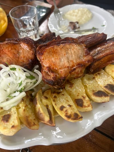 A platter of grilled pork chops and ribs, accompanied by potato wedges, sliced onions, and chopped herbs, sits on a wooden table. To the side, there's a plate with mashed potatoes and a fork. A glass and a lemon wedge also appear near the platter. The food is presented in a rustic setting, highlighting the charred texture of the meat and the simple accompaniments.