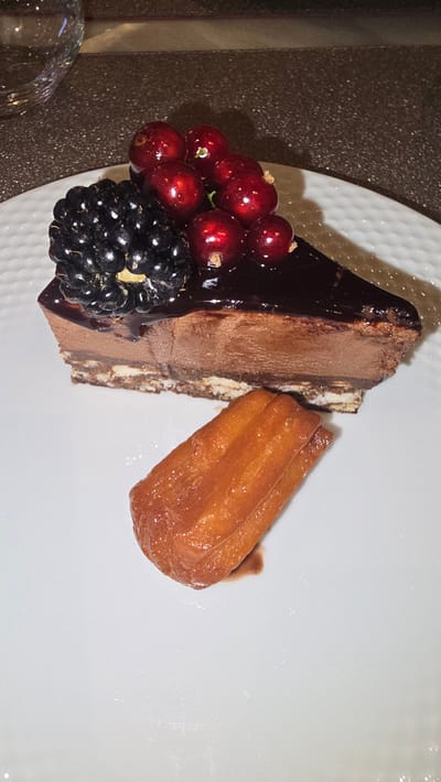 A close-up, top-down shot displays a slice of chocolate cake on a white plate. The cake has multiple layers, with a dark chocolate glaze on top, and is garnished with a cluster of red currants and a blackberry. To the right of the cake, a single golden-brown pastry, possibly a churro or a similar fried dough confection, sits on the plate with a small pool of sauce beneath it. The background is a textured, dark surface, suggesting a restaurant or cafe setting. The lighting in the image is somewhat dim, with a slight sheen on the dessert items, indicating a possible indoor setting. There is no visible text in the image. The overall presentation suggests a dessert course being served. The angle and composition focus solely on the food.