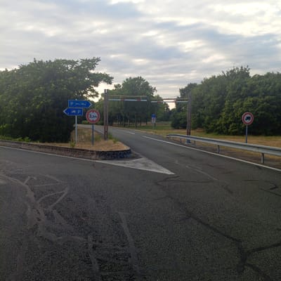 The image depicts a road junction with signage and trees in the background. On the left, there are two blue directional signs. The top sign points right and shows a parking symbol with icons for a car, a caravan, and a truck. The bottom sign points left and also shows a parking symbol with icons for a car and a caravan. To the right of these signs is a circular red and white sign with "3,3m" inside, indicating a height restriction. Further to the right, on the road, is a barrier with red and white stripes. Another circular sign on the right side of the road shows a truck symbol within a red circle and a diagonal red line, signifying a prohibition for trucks. The road surface is asphalt, with some tire marks visible on the left side. The environment is characterized by lush green trees on either side of the road and a cloudy sky. The location is identified as Saint-Augustin-des-Bois, France.