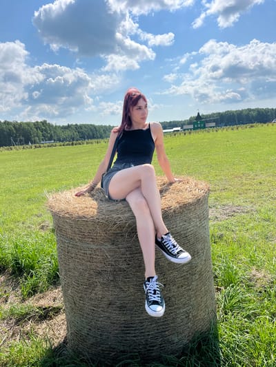 A young woman with reddish-brown hair is sitting on a large, round bale of hay in a grassy field. She is wearing a black tank top, denim shorts, and black Converse sneakers. Her legs are crossed, and she is looking off to the right side of the frame. The background shows a green field, a line of trees, and a few buildings in the distance. The sky is blue with white, fluffy clouds. The location is the city of Kurtsevo (Kovriginskii S/S), Russia.