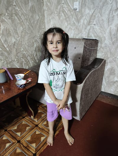 A young girl stands in a room with patterned wallpaper and a tiled floor. She is wearing a white t-shirt with a graphic of mountains and trees, and purple shorts with a floral pattern. Her hair is dark and pulled into two pigtails. She is standing in front of a brown and beige armchair. To her left is a wooden table with a bowl and a container of what appears to be yogurt, along with other small items. The room appears to be a home interior, possibly in the city of Navruz, Uzbekistan, given the context. The lighting suggests an indoor setting, possibly during the evening or in a room with artificial light.
