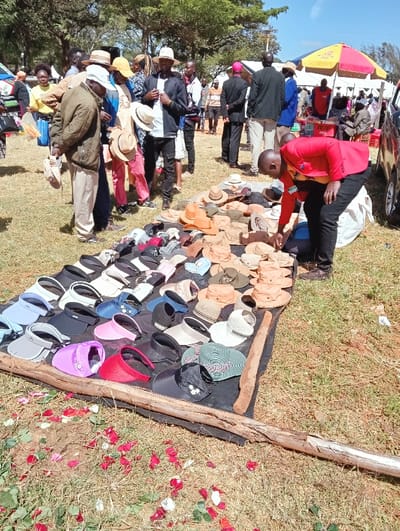 A bustling outdoor market showcases a wide array of hats and visors laid out on a black tarp. Customers browse the selection, including fedoras and sun visors in various colors and styles. A vendor in a red jacket assists a customer. People mill about in the background, and a tent is visible, suggesting a festival or market event. Scattered rose petals add a touch of festivity to the scene.