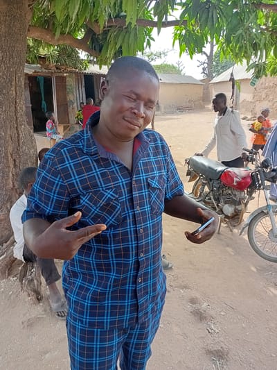 The image captures a man in Nata-Ala, Nigeria. He is wearing a blue plaid shirt and standing outside. Behind him, there's a motorcycle, another man in a white shirt, and a few buildings. A tree is on the left side of the frame. The ground appears to be dirt or sand.