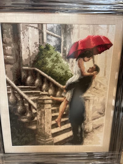 The image displays a framed cross-stitch artwork depicting an outdoor urban scene in Disevitsa, Bulgaria. Two individuals are centered in an intimate embrace. The male figure, dressed in dark clothing, is actively lifting the female figure off the ground. The female, wearing a light-colored sleeveless garment, has her right leg exposed and bent, with her bare foot visible. She holds an open red umbrella directly over both figures. Her arms are wrapped around the male's neck, and their faces are in close proximity, indicating a kiss or deep embrace. They are situated on a stone staircase, characterized by multiple tiers and a balustrade featuring ornate balusters. To the left of the figures, a dense volume of green foliage is present. The background consists of textured architectural elements, consistent with stone or concrete construction. The cross-stitch is enclosed within a dark frame and a lighter mat, all covered by clear plastic film, which introduces some glare and distortion to the image.