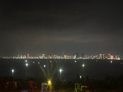 The scene is a nighttime panorama of the Da Nang city skyline viewed from Sơn Trà Ward, Vietnam. The foreground features dark ground cover and several plastic chairs of red, blue, and green coloration. Two yellow-glowing lanterns are visible on the ground, producing prominent starburst lens flares. The midground consists of a dark body of water, likely a bay or river, reflecting numerous bright, starburst-shaped lights. The background displays a densely illuminated city skyline. High-rise buildings with varying light patterns are visible, including a central skyscraper emitting a distinct red illumination and others featuring white, yellow, and blue lights. The sky is dark and overcast, showing no celestial bodies. Faint, elongated light streaks are present in the upper-center sky. No individuals are visible.