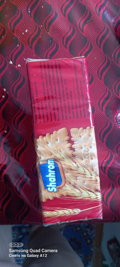 The image shows a close-up of a package of "Shahrom" brand crackers, possibly in Levakant, Tajikistan. The packaging is red and features an illustration of wheat stalks and the brand name in a blue and orange font. Text in a foreign language, likely Tajik or Russian, is printed on the packaging, providing information such as ingredients, production date, and expiration date. The text also includes contact information, with a phone number starting with "+992", a country code for Tajikistan. The crackers themselves are visible through the transparent part of the packaging, appearing square-shaped and light brown in color. The background is a red and black patterned fabric, possibly a tablecloth or a decorative surface. The lighting suggests it might be indoors, possibly during the day. The overall impression is a product shot, focusing on the food item and its packaging.