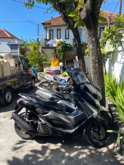In Denpasar Barat, Indonesia, a sunny day is depicted with a street scene featuring multiple motorcycles parked under trees. In the foreground, two dark-colored scooters are parked side-by-side, with the closest one being a Yamaha NMAX. Behind them, a green and white dirt bike is visible, followed by another motorcycle. A small black utility truck is parked to the left, loaded with light-colored boxes. In the background, traditional Balinese architecture with red-tiled roofs and white walls is visible, along with lush green vegetation and palm trees. A religious offering, possibly a Balinese ceremony, is set up with a yellow cloth and a white umbrella. The environment appears to be residential or commercial, with power lines visible overhead. The overall impression is of a typical day in a tropical urban environment.