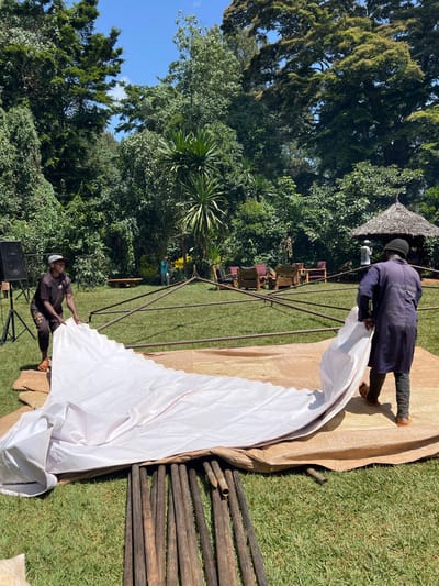 In an outdoor setting in Mururiini, Kenya, two men are actively engaged in setting up a large white tent or canopy. The scene takes place on a bright, sunny day, indicated by the clear sky and visible shadows on the green grass.
One man, dressed in a dark blue long-sleeved tunic with white text on the back (partially legible, possibly including "EVENT"), dark trousers, and sandals, is positioned towards the right side of the frame. He is holding and spreading out a large, white fabric, which appears to be the main covering for the tent. Underneath this white fabric, a larger tan-colored tarpaulin is laid out on the grass.
Towards the lower-left, a second man wearing a dark t-shirt, dark pants, and a white baseball cap is crouched or standing near a speaker on a stand, also handling the white fabric and possibly connecting it to the metal frame of the tent. A structure of metal poles, forming part of the tent's framework, is visible between the two men. To the far right, a stack of wooden poles lies on the grass, likely intended for the tent's construction or another nearby structure.
In the background, lush green trees and tropical-looking foliage dominate the landscape. Several wooden chairs with reddish cushions are arranged in rows, suggesting a prepared seating area for an event. A small, round thatched structure can also be seen in the upper-center background, nestled among the trees. The overall impression is one of preparations for an outdoor gathering or function.