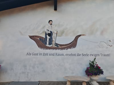 This image shows a mural painted on a white wall, located in Schwangau, Germany. The mural depicts a man in historical attire standing in a boat. He is wearing a white robe with blue trim, a dark hat, and holds a staff in his right hand and a hat in his left. The boat is being pulled by two white swans. Below the mural, text is inscribed in German, reading "Als Gast in Zeit und Raum, ersehnt die Seele ewigen Traum!". To the right of the text, there is a large urn filled with colorful flowers, and two stone benches are visible in the foreground.
