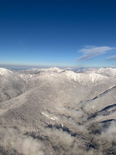 A landscape features a clear, blue sky transitioning into a misty horizon. Mountain ranges, covered in snow, dominate the scene. Patches of clouds or mist cling to the mountainsides, creating textured patterns. The overall image suggests a high-altitude view over a wintery, mountainous region.