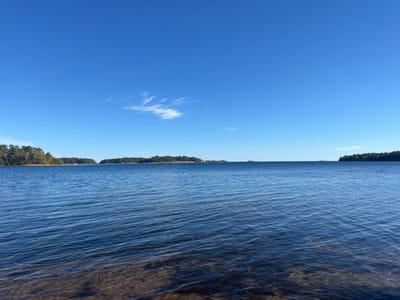 A wide-angle, static image captures a tranquil marine landscape within the Västervik archipelago, Västervik, Sweden. The lower third of the frame shows calm, clear blue water, with the shallow foreground revealing a light-colored, presumably sandy or rocky, bottom. The midground consists of a deeper expanse of dark blue water extending towards the horizon. In the distance, several low-lying landmasses, appearing as islands or distant shorelines, are visible, densely covered with green trees. The upper two-thirds of the image display a clear, vivid blue sky, punctuated by a single, elongated wispy white cloud in the upper-left sector. No human figures, animals, man-made structures, or activities are depicted. The scene suggests an unobstructed natural environment under bright daylight conditions.