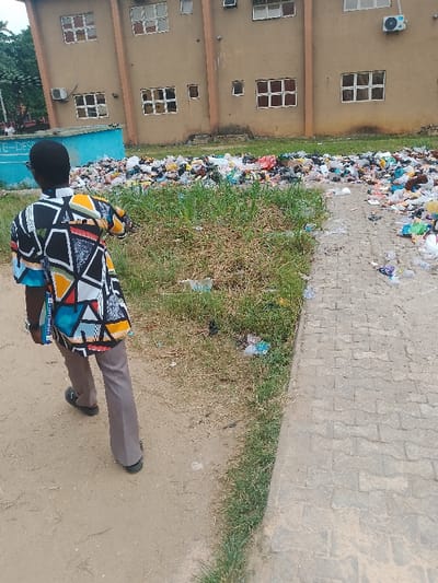 An outdoor scene in Rumuekini, Nigeria, shows an individual walking on a dirt path adjacent to a large accumulation of refuse. The subject, viewed from the rear, is a person wearing a short-sleeved, multi-patterned tunic in black, white, yellow, blue, red, and green, grey trousers, and dark open-toed footwear. A blue and white lanyard, partially displaying "I LOVE UNIB...", is visible on the subject's left side.
To the subject's right, a narrow strip of sparse grass borders the dirt path, which then transitions into a paved walkway. Both the grassy area and the walkway contain scattered pieces of refuse. Immediately beyond the grassy strip and extending along the base of a two-story, light brown building, is a substantial mound of mixed refuse, primarily composed of plastic bags, bottles, and other discarded items. The building features multiple rectangular windows with either white or red frames and several wall-mounted air conditioning units. To the left of the refuse pile, a light blue structure with the partial inscription "E-DES" is visible. In the distant background, trees and the faint outlines of other individuals are present under an overcast sky.