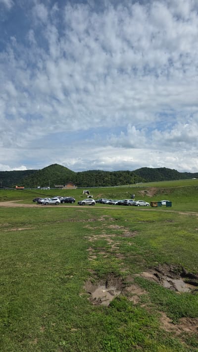 The image captures a pastoral landscape with a cloudy sky and rolling green hills in the background. A line of parked cars is visible on a dirt and grass area in the mid-ground. Beyond the cars, a road runs horizontally with a few buildings and structures along its side, including what appears to be a small warehouse or barn. The foreground features a patch of muddy ground with visible tire tracks and standing water. The overall scene suggests a rural or recreational area in the vicinity of Таш-Асты, Russia, with elements of both natural scenery and human activity.