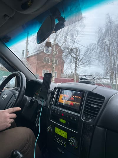 The image depicts the interior of a vehicle, looking forward through the windshield towards an exterior environment identified as Yasenevo, Russia. A person, likely the driver, has their right hand positioned on a black Kia steering wheel, displaying a gold ring on the ring finger. The driver's lower body is clad in light brown trousers. Mounted on the left side of the vehicle's center console is a smartphone, vertically oriented and connected to a light blue charging cable. The center console features an infotainment screen displaying a still image with predominant red and brown tones, possibly depicting food or embers. Below the screen are various control buttons, including a triangular red hazard light button, and a digital climate control panel indicating a "26.0" degree setting for dual zones. A set of car keys hangs from the ignition. A small, black model helicopter is placed on the dashboard. The windshield's upper section has a blue tint, and a rearview mirror is centrally affixed. A square, clear air freshener bottle with a wooden top hangs from the rearview mirror, next to a mounted dashcam. The exterior scene, viewed through the windshield, shows an unpaved or dirt road in the foreground with traces of snow or slush. A prominent two-story red brick house with multiple windows stands in the middle ground, behind a utility pole. The background is populated by numerous deciduous trees with bare branches under an overcast sky.