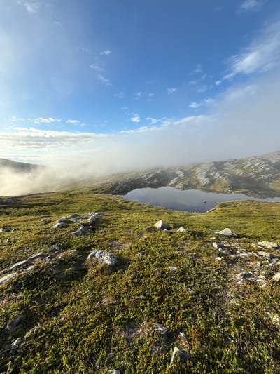 The image depicts a panoramic view of a rugged, mountainous landscape in Aun, Norway, during what appears to be either early morning or late evening, given the soft lighting and mist. The foreground is covered in low-lying green vegetation and scattered grey rocks. In the mid-ground, a calm, dark body of water, likely a mountain tarn or small lake, reflects the sky and surrounding terrain. The water's edge is bordered by rocky outcrops and more of the dense green ground cover. Further into the background, a thick layer of mist partially obscures rolling hills and distant mountains, creating a sense of depth and mystery. The sky above is a clear blue with scattered white clouds, some of which are tinged with sunlight. The overall impression is one of serene, untamed natural beauty.