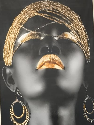 The image is a close-up, low-angle, black and white photograph of a person's face from the neck up. The person has dark skin and is adorned with elaborate gold jewelry. A thick band of gold beads is worn across the forehead and draped down the sides of the head. Gold makeup is applied to the eyelids, and the lips are painted a vibrant metallic gold with a textured finish. The person is wearing large hoop earrings, also decorated with gold beads and metallic accents. The background is uniformly black, isolating the subject and highlighting the intricate details of the jewelry and makeup. There is no indication of the city of Константиновка, Russia, in the image itself.