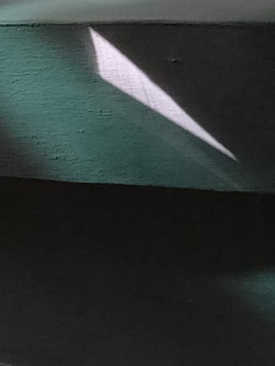 The image depicts a close-up view of a textured, dark green surface with a sharp, bright white triangular light beam cutting across it from the upper left to the lower right. The surface appears to be a wall or a similar flat structure, possibly made of concrete or stucco, exhibiting a slightly rough texture. The light beam is well-defined, suggesting it is cast by a window or a similar opening. The background is mostly dark, with the lower portion of the image being entirely black, implying a dimly lit environment. The upper edge of the green surface has a lighter, smoother band. The light beam appears to be the sole subject of interest, creating a stark contrast against the dark background and the textured surface. The time of day is suggested by the sharp, defined light, possibly late afternoon or early morning, or indoor lighting. There is no text visible in the image, nor are there any people, animals, or other objects. The overall mood is one of simplicity and focus on light and shadow. The provided location context of Ternovka, Moldova, does not offer specific visual cues within the image itself but suggests the geographical origin of the photograph.