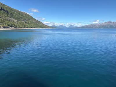This media file depicts a scenic landscape of a body of water with mountains in the background. The scene is set on a clear, sunny day, with blue skies and scattered white clouds. The foreground is dominated by the calm, blue water, with subtle ripples on its surface.
To the left, a lush green, tree-covered mountainside slopes down towards the water. The mountains in the distance are varied in shape and size, with some appearing rocky and others with hints of snow or vegetation. The overall impression is one of natural beauty and tranquility.
The image appears to be taken from a vantage point near the water's edge, possibly a dock or shoreline. There are no people or specific objects prominently featured, emphasizing the natural elements of the scene. The time of day is likely daytime, given the bright sunlight and clear skies. The location cues, such as the type of mountains and the presence of a fjord-like body of water, are consistent with a coastal or mountainous region.
The general mood evoked is one of peace, serenity, and awe inspired by the vastness of nature. There is no visible text within the image.