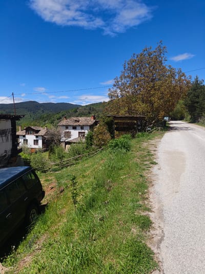 The image depicts a rural scene in Kovachevitsa, Bulgaria. A narrow asphalt road curves through the landscape. To the left of the road, there is a grassy verge with vegetation. Partially visible on the left is a dark-colored SUV parked on the incline. Traditional houses with white walls and dark tiled roofs are situated further back, nestled among trees and greenery. In the background, there are forested hills under a blue sky with scattered clouds. A large tree with yellow leaves stands near a wooden structure (possibly a sign) along the roadside. Power lines stretch across the sky.