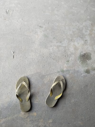 This media file shows a pair of light brown, textured flip-flops resting on a concrete surface. The flip-flops are positioned on the lower left and lower right portions of the frame, with a significant amount of empty concrete space above them. The concrete appears somewhat weathered, with a few subtle marks and variations in shade, suggesting an outdoor setting. The lighting indicates it is daytime, with soft shadows cast by the flip-flops. There is no discernible activity taking place; the flip-flops are stationary. The text "SWALLOW" is visible on the strap of the left flip-flop. The overall impression is one of simplicity and everyday objects in a common environment.