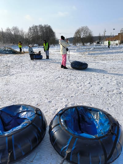 The image depicts an outdoor, snow-covered scene in Prybuzke, Ukraine, under a clear blue sky. In the immediate foreground, two black and blue inflatable snow tubes are visible. The midground features several individuals and objects: a person wearing a white jacket, pink pants, and a black hat stands, holding a smartphone in a horizontal orientation, appearing to take a photograph. An inflatable snow tube rests on the snow near this individual. Further back, a child is seated in a dark blue inflatable snow tube, attended by a person in a light-colored jacket. Nearby, another individual in a green high-visibility vest and dark pants stands. To the far left, a person in a yellow high-visibility vest is partially visible near a mound covered with a tarp, holding a shovel. To the far right, another person in a green high-visibility vest is also seen holding a shovel. The ground is covered in white snow with visible tracks and undisturbed areas. The background consists of a treeline with bare deciduous trees, utility poles, and distant structures, possibly residential or agricultural, under bright sunlight.