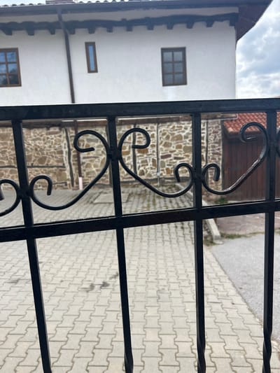 The image depicts an exterior scene in Gigintsi, Bulgaria, viewed through a wrought iron gate. The gate, positioned in the foreground, is black with decorative scrolled metalwork forming heart-like shapes and circular motifs. Beyond the gate, a light grey interlocking paver driveway or courtyard occupies the midground, exhibiting some dark, irregular spots.
Behind the paved area, a low stone wall constructed from varying sizes and colors of natural stone, interspersed with vertical wooden beams, serves as a boundary. To the right of the stone wall, a dark brown wooden plank door is visible under a terracotta-tiled roof section.
In the background, a two-story building with white stucco walls is prominent. It features dark brown wooden window frames; one on the left has a multi-pane grid design, and another on the upper floor is a narrow, vertical single-pane window. A brown gutter downspout is attached to the building's facade. Dark wooden eaves are visible under the roofline. The sky above is partially cloudy with grey and white formations. No individuals or specific actions are depicted within the frame.