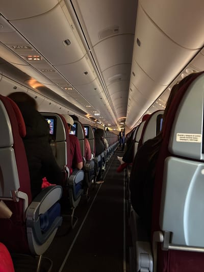 The image depicts the interior of an aircraft cabin during flight. Rows of maroon-colored passenger seats with grey and blue headrests are visible, occupied by seated individuals who are either asleep or watching personal screens on the seatbacks in front of them. Overhead compartments line the left side of the image. The lighting is dim, and the overall atmosphere suggests a nighttime flight.

There is no information within the image to suggest any relation to the city of Pudahuel, Chile.