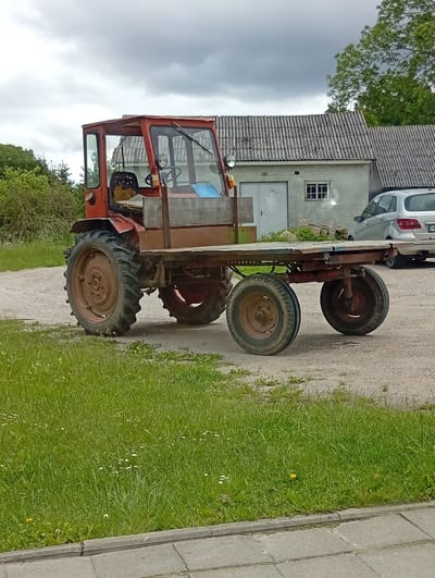 An outdoor scene in Šaukėnai, Lithuania, features a red tractor with a flatbed trailer attached, parked on a gravel surface. The tractor has a cab with windows, a steering wheel visible inside, and a driver's seat. The trailer is empty and appears to be made of wood and metal. In the background, a light-colored, multi-story building with a corrugated roof stands, and a silver car is parked to the right of the building. Green grass with scattered white flowers lines the foreground and the left side of the scene. The sky is overcast with dark clouds.