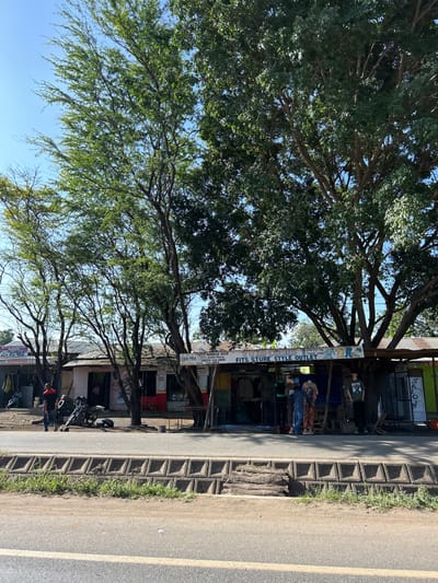 This image depicts a vibrant roadside scene in Mto Wa Mbu, Tanzania, under a clear blue sky. Large, mature trees with dense green foliage dominate the upper and central parts of the frame, providing ample shade. Below the canopy of trees, a series of single-story shops or businesses line the street.
A paved road runs along the right side of the image, separated from the commercial establishments by a distinctive patterned concrete sidewalk or curb. Several people are visible near the shops; one person in a blue shirt stands in front of a storefront displaying goods, another in a green shirt is further back, and a person in a red hat is near a parked motorcycle. The shops feature corrugated metal roofs and open fronts, showcasing various items, including clothing.
A prominent white banner is stretched between two trees, advertising a business. The text on the banner reads: "FITS STORE STYLE OUTLET". Below this, partial text includes "TEL: 0753 913...", "TEL: 0767 77...", "WE DEAL IN...", and "JUMBA LA WENYE...". Another partially visible sign on a nearby building indicates a "SALON".
The bright sunlight and clear sky suggest it is daytime, likely morning or early afternoon, indicating good weather. The scene captures the everyday activity of a market town, with shops open for business and people moving casually.