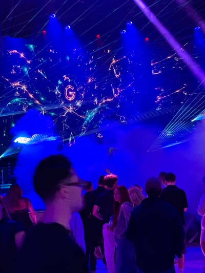 The image depicts a dimly lit indoor scene, possibly a nightclub or concert venue, with a strong blue and purple lighting scheme. The background features a large screen displaying an abstract, glowing pattern with a prominent circular "G" logo. Laser lights crisscross the space, adding to the dynamic atmosphere. Smoke or haze is present, enhancing the effect of the lights. In the foreground and mid-ground, several individuals are visible, mostly seen from the back or side. They appear to be standing, suggesting they are part of a crowd or audience. Some are dressed casually. The overall impression is one of a lively, modern entertainment event. The specific location of Akhalubani, Georgia, is not discernible from the visual information provided in the image.