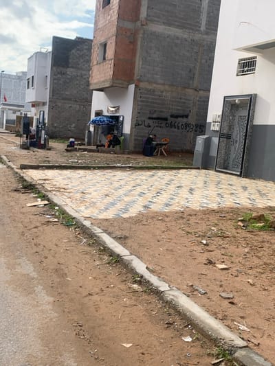 This image depicts a street scene in Ameur عامر, Morocco, under an overcast sky. The foreground features a dirt road with a damaged concrete curb separating it from an unkempt dirt shoulder containing scattered debris. Beyond the curb, a paved area with light and dark grey diagonally striped pavers extends towards three multi-story buildings.
From left to right, the buildings are:
1. A partial view of a white, finished structure with a visible Moroccan flag pole and a red flag. Adjacent to this, two red and grey fuel pumps are present.
2. A partially constructed building, featuring a finished white ground floor with a dark blue awning and a blue-and-white striped umbrella. This area functions as an outdoor seating space, where two individuals are seated at a small table. One individual wears a dark top, the other an orange-brown top. The upper floors of this building consist of exposed grey concrete blocks and reddish-brown bricks with rebar visible, indicating ongoing or incomplete construction. Graffiti, including Arabic text and a phone number (0666089385), is present on the grey concrete wall. Further to the right of the umbrella, a third individual in a dark jacket sits at a small orange table with a white plastic chair, engaged with an object on the table. A dark blue container sits next to this individual.
3. A finished white building with a grey base. A large, ornate, dark-colored metal door frame leans against the white wall. A window with metal security bars is visible on an upper floor.
The overall environment suggests a mix of residential and small-scale commercial activity, with construction in progress or stalled in the vicinity.