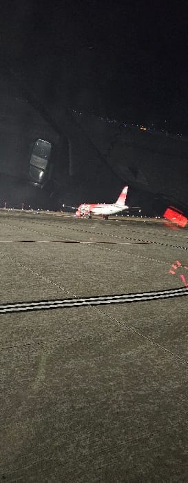The scene depicts an airport tarmac at night, specifically at Katowice International Airport (KTW) in Pyrzowice, Poland. A white passenger jet aircraft with prominent red markings on its tail and fuselage is positioned on the paved surface, likely an apron or taxiway. The aircraft appears to be an Airbus A320 series. Numerous small, illuminated airport lights, possibly taxiway edge lights, are visible in the mid-ground, extending into the distance. To the far right, a large, bright red object, possibly ground support equipment or a barrier, is partially visible. The foreground features a dark, concrete or asphalt surface marked with white dashed lines and solid black lines, indicative of airport ground markings. The image is captured from a slightly elevated perspective, with a dark aircraft wing partially visible in the upper left quadrant of the frame, showing some reflective elements. No individuals are discernible. The overall environment is dark, illuminated by artificial airport lighting.