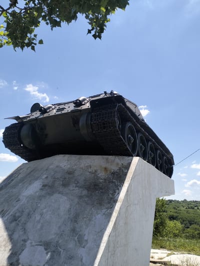 The image shows a close-up, low-angle view of a historical tank monument situated on a concrete pedestal. The tank, which appears to be a Soviet-era model, is dark green and shows signs of wear. Its tracks and road wheels are clearly visible. The pedestal is made of rough, weathered concrete with some cracks and discolorations. The monument is located outdoors, with a bright blue sky with scattered white clouds in the background. Green foliage from a tree hangs into the upper left corner of the frame, and a lush, green, tree-covered hillside is visible in the background to the right, suggesting a rural or park-like setting. The overall scene is one of outdoor monumentality and historical display. This monument is located in the village of Cinișeuți, Florești District, Moldova.