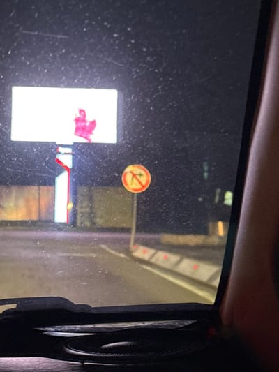 View from inside a vehicle with a dirty window. In the foreground, a "no loitering" sticker is visible, along with a display screen showing a white rectangle with some red coloration. The exterior shows a partial building, with red lights. A partially visible rear-view mirror is to the right.