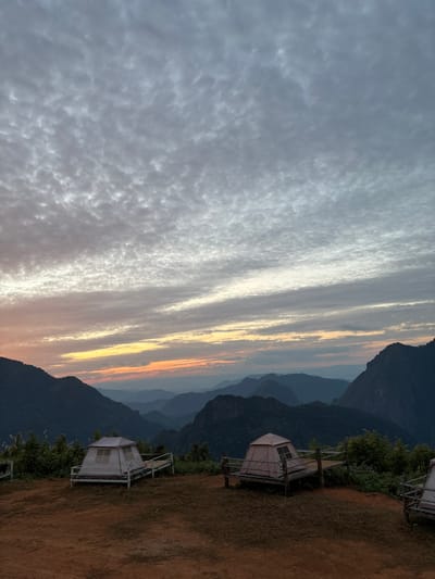 The image displays a mountainous landscape at either sunrise or sunset in Nong Ian, Laos. The sky is dominated by an extensive altocumulus cloud formation, appearing grey and white, with distinct horizontal bands of orange, yellow, and pink light near the horizon. Multiple layers of forested mountains recede into the distant background, showing increasing blue atmospheric haze. The immediate foreground consists of a reddish-brown dirt and sparse grass expanse. On this elevated ground, two dome-style tents are erected on distinct, elevated wooden platforms featuring low railings. The tent on the left is light-colored and sits on a white-framed platform. The tent on the central right is beige and rests on a dark-framed wooden platform. A third, partial platform is visible on the far right. No individuals or animals are present in the scene.
