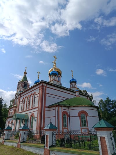 A low-angle, full shot captures the facade of a white brick church with red trim and a green roof in Staryi Pokrov, Russia. Several golden-domed cupolas with blue accents and crosses rise above the main structure. A bell tower with a green, multi-tiered roof is visible to the left, also topped with a cross. A black metal fence with ornate designs and white brick pillars encloses the churchyard. The sky above is a bright blue with scattered white clouds. Trees can be seen behind the church and to the right. The overall scene depicts a religious building on a sunny day.