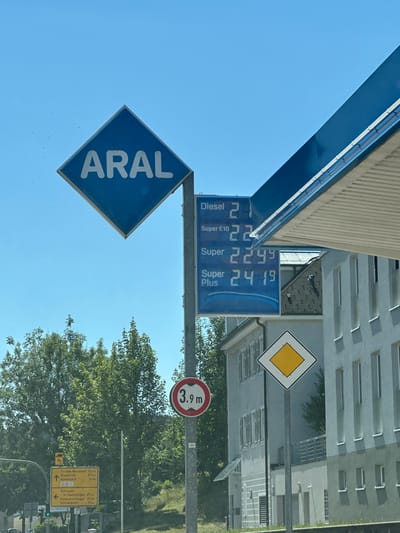 The image shows a gas station sign with prices for different types of fuel. The sign displays "ARAL" in large white letters on a blue diamond shape. Below it, a blue rectangular sign lists fuel types and prices: "Diesel 2.1", "Super E10 2.2", "Super 2.299", and "Super Plus 2.419". To the right of the fuel price sign, a section of the blue gas station canopy is visible. In the background, a modern building with many windows stands under a clear blue sky. To the left, a road with trees and directional signs is visible. One sign indicates distances to nearby towns, including Titisee-Neustadt, Waldkirch, and Gutenbach. Another sign points towards Villingen, with distances to Schwenningen and Donaueschingen. A circular traffic sign indicating a height limit of 3.9 meters is also present. A yellow diamond-shaped sign, typically indicating a priority road, is mounted on a pole in front of the building. The overall scene suggests a typical roadside view of a gas station in a German town.