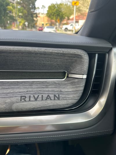 A close-up interior shot displays a section of the dashboard. The word "RIVIAN" is boldly engraved vertically on a gray wood-grain panel. A long, textured speaker grill runs parallel to the name. Above the speaker, a metallic accent bar reads "UNTRODDEN, FOREVER." The panel is framed by a silver metallic trim and dark leather. An air vent is partially visible above the panel, with hints of a car window and exterior foliage in the background.