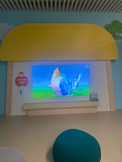 In Hwaseong-si, South Korea, a digital display is mounted on a wall, simulating a thatched roof. The display shows an animated character, possibly an ice cream cone, with large eyes and a purple body, interacting with blue crystalline structures against a vibrant blue and green background. To the left of the screen, a red circular emblem with Korean text and a smaller circular button are visible. The display is positioned above a light-colored wooden shelf. On the shelf to the right of the screen, a small book or pamphlet with an image of people on it rests in a triangular stand. The surrounding environment appears to be an indoor educational or entertainment space, with light-colored walls and a beige floor. Two round, soft seating elements, one light gray and one teal, are visible in the foreground.