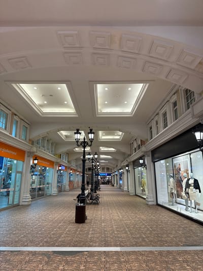 An indoor hallway is lined with store windows and punctuated by old-fashioned streetlights with benches. The ceiling is high, white, and ornate, featuring arched structural elements with decorative square panels and recessed lighting. The floor is paved with small, geometric tiles. The hallway seems to stretch a considerable distance, with diminishing perspective suggesting a long corridor of shops and amenities.