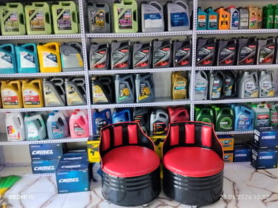 The image depicts an indoor scene, likely a store or workshop, filled with shelves stacked with various automotive fluids and products. The shelves are organized and filled with numerous bottles and containers of motor oil, antifreeze, and other automotive fluids, all labeled with different brands and specifications. In the foreground, two unique barrel-shaped chairs with red seats and black padded backs are positioned. These chairs are placed on a marble-patterned floor. The overall impression is one of an organized retail space for automotive supplies. The location of Yovon, Tajikistan is mentioned, but there are no specific visual cues in the image to confirm or deny this location. There are no people visible in the image.