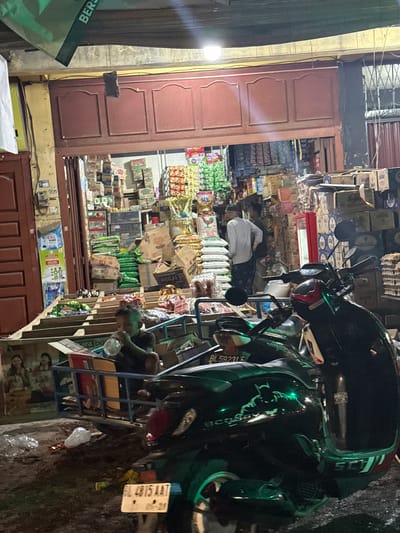 The media file depicts a nighttime scene outside a small shop in Lampeuneurutblang, Indonesia. A dark-colored motorcycle, possibly a scooter, is parked prominently in the foreground, facing towards the right of the frame. It appears to be a green and black model, with visible decals and a license plate reading "BL 5923 AAT" and "05-25".

Behind the motorcycle, the shop's interior is visible, filled with various products stacked on shelves. These appear to be food items, drinks, and other packaged goods, with many cardboard boxes and plastic packaging. The shop is lit by artificial lights, casting a warm glow on the merchandise.

A person wearing a light-colored shirt is partially visible inside the shop, appearing to be engaged in some activity related to the products. Another person is partially visible near the bottom right of the frame. The overall atmosphere suggests a busy local store, possibly a convenience store or a small grocery.

The ground in front of the shop appears to be a dark, possibly wet, street or pathway. There are some discarded plastic bags and debris on the ground, indicating a typical urban street environment. The scene is captured at night, as suggested by the artificial lighting and the dark sky. The building facade features traditional wooden doors painted a reddish-brown color. A fluorescent lamp is visible on the left side, illuminating the entrance area.
