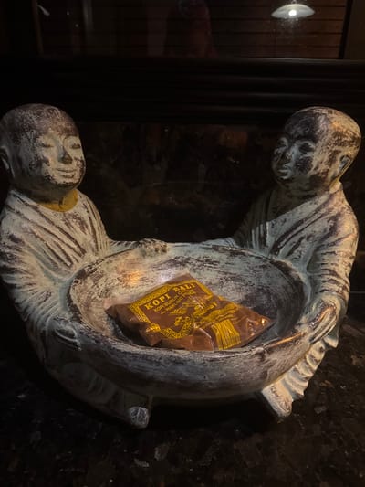 An ornate statuette featuring two weathered, child-like figures, resembling monks, sits on a dark, speckled surface in Tibubiyu, Indonesia. The figures, rendered in a pale, aged material with brown and green undertones, face each other, jointly supporting a large, shallow bowl between them. The figure on the left has closed eyes and a slight smile, while the figure on the right has open eyes. A single brown plastic packet, labeled 'KOPI BALI' and 'KOPI BUBUK CAP WARUNG POJOK' in yellow text, rests inside the bowl. The background is dark and reflective, showing indistinct reflections of a light source and a reddish form.