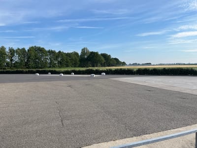 The image depicts an outdoor asphalt surface, likely a parking lot or road, in Oosterland, Netherlands. Several white, rounded bollards are visible in a line along the edge of the asphalt, near a hedgerow. Beyond the hedgerow, there is a field of crops, possibly wheat or barley, stretching towards a distant treeline. The sky is blue with scattered wispy clouds. A light-colored concrete area borders the asphalt in the background. The overall scene is rural and agricultural, with no people or vehicles present.