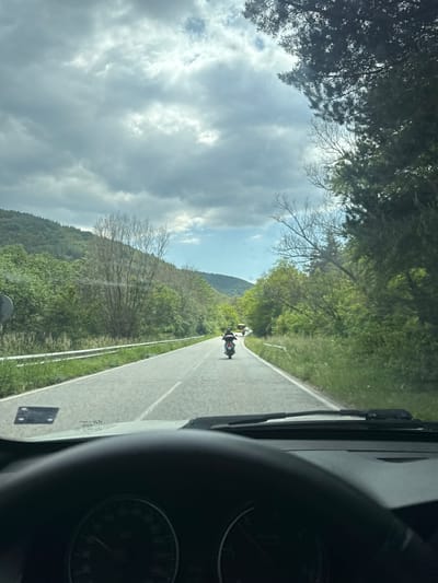 The image presents a forward-looking view from inside a vehicle, likely a car, traveling on a road through a rural, hilly landscape in Cherna Mesta, Bulgaria. Part of the vehicle's dashboard and the rim of a steering wheel are visible on the right side of the frame, with the windshield and a wiper arm on the left.
Ahead on the asphalt road, two motorcycles are visible, moving in the same direction as the observer. The closer motorcycle appears dark, while the one further ahead is lighter in color. The road features a clear white center line and a white line marking its right shoulder. A metal guardrail runs parallel to the road on the right, separating it from the sloped terrain.
The surrounding environment consists of rolling hills heavily covered in a mix of green deciduous trees and some coniferous trees, alongside scattered bare-branched trees, suggesting early spring foliage. The sky above is largely overcast with prominent, bright clouds, indicating daytime. The overall scene depicts a journey or scenic drive through a natural, forested area. No distinct text is clearly legible on any objects within the image.