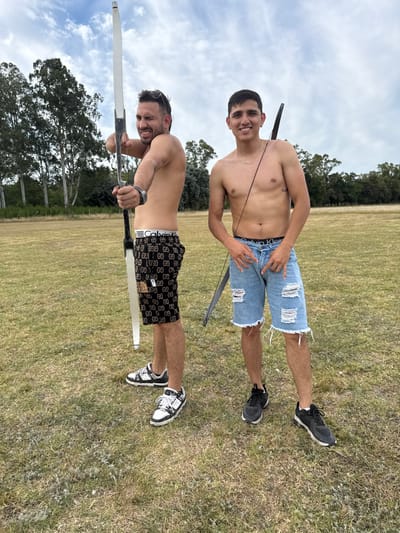 Two shirtless men are in a grassy field, each holding a bow. The man at the bottom of the frame is pulling back on his bow, seemingly aiming, wearing dark shorts and white sneakers. The other man, above him, has a bow pointed upwards, wearing denim shorts and dark sneakers. The men are both smiling. Trees and sky are visible in the background.
