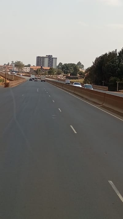 This image depicts an elevated multi-lane asphalt highway in Gitaru ward, Kenya, under a bright, largely clear sky. The dark grey road surface is marked with white dashed lane lines, indicating multiple lanes of traffic. Concrete barriers line both sides of the elevated roadway structure. On the far left, an exit or on-ramp structure is visible. Vehicles are present in both directions of travel on the highway. On the left-side lanes, multiple vehicles, including a light-colored truck and several cars, are in motion, traveling away from the camera's perspective. On the right-side lanes, a white pickup truck, a dark sedan, and another dark vehicle are in motion, traveling in the same general direction as the camera's perspective. A motorcycle is discernible further down the right side between the concrete barriers. The background on the left features a collection of buildings, including a prominent multi-story high-rise structure with a dark facade, interspersed with deciduous and evergreen trees. The background on the right is dominated by a dense growth of evergreen trees. No individuals are visible within the frame.