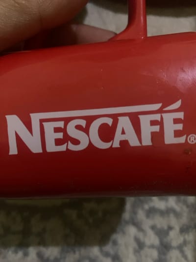 A close-up shot focuses on a red mug prominently displaying the white "NESCAFÉ" logo. The brand name is vertically oriented along the mug's body, with the handle visible on the left. A hand partially holds the mug. The background presents a blurred surface with a mottled gray and white pattern. The image has an informal, snapshot aesthetic.